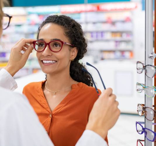 African young woman in optic store choosing new cool-down glasses with optician. Mixed race girl trying new eyeglasses with the help of the pharmacist. Happy multiethnic woman trying new rest spectacle frame in optic store: eyesight and eye care concept.